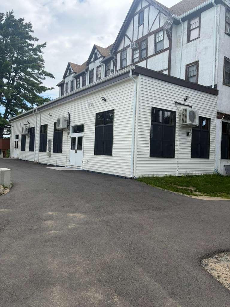 A single-storey hall extension built onto an existing gabled building, clad in white siding with tall windows down its length and a pair of doors onto the drive