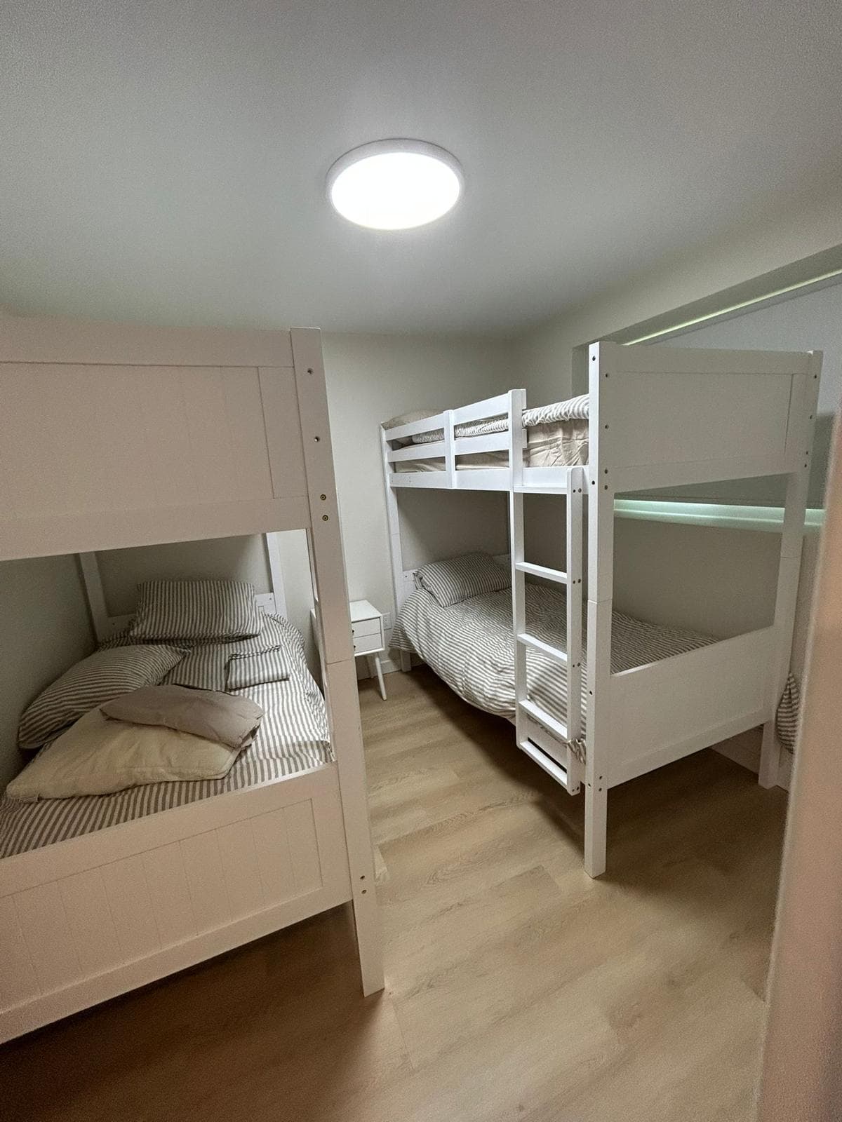 A bunk room with two white timber bunk beds made up along facing walls, a small nightstand between them and plank flooring run through