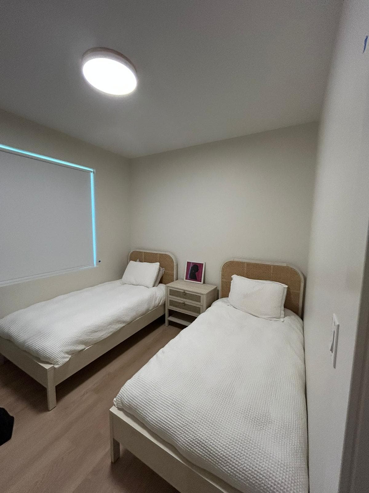 A finished sleeping room with two single beds under cane headboards, a nightstand between them and a wide window along the far wall