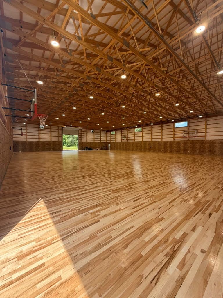 The finished basketball court inside a camp gym — a laid hardwood floor running the full clear span under exposed timber roof trusses