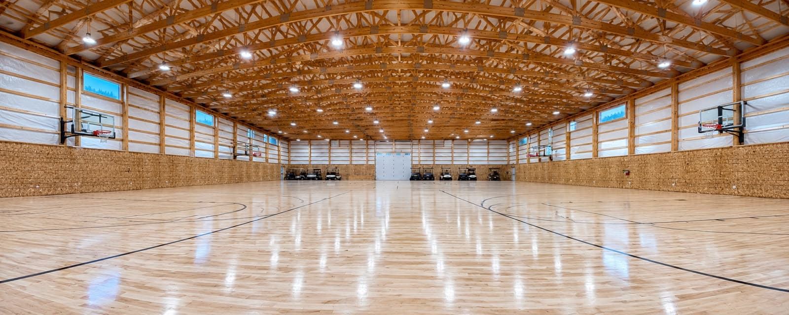 A finished gym with two full basketball courts on one hardwood floor, hoops along both side walls and timber trusses spanning the whole hall overhead