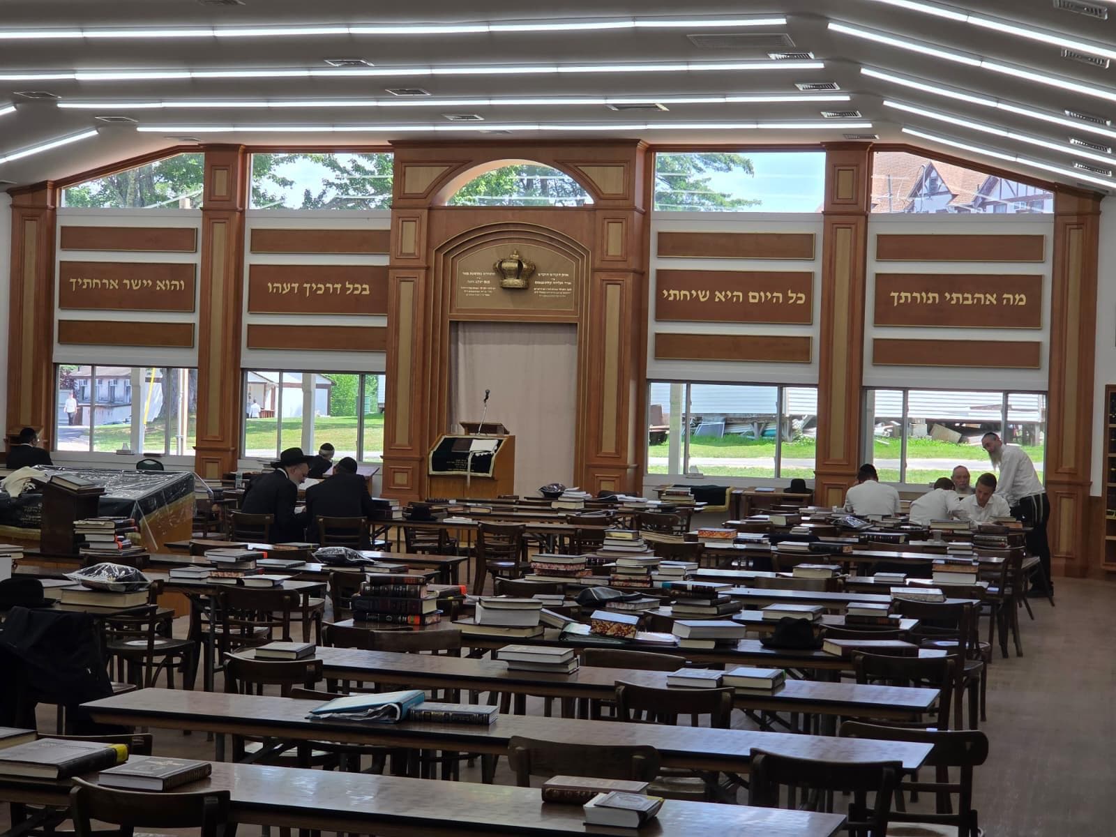 The front of a beis medrash in daylight — a panelled timber wall with lettered panels either side of the aron kodesh and an amud below it, full-height windows down both side walls