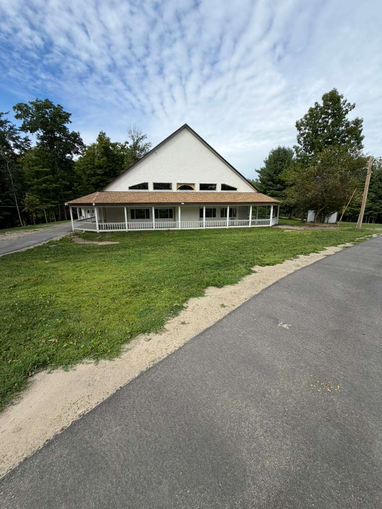 The front elevation of a shul across mown ground, its steep hipped roof rising to a band of clerestory windows above a full-width covered porch