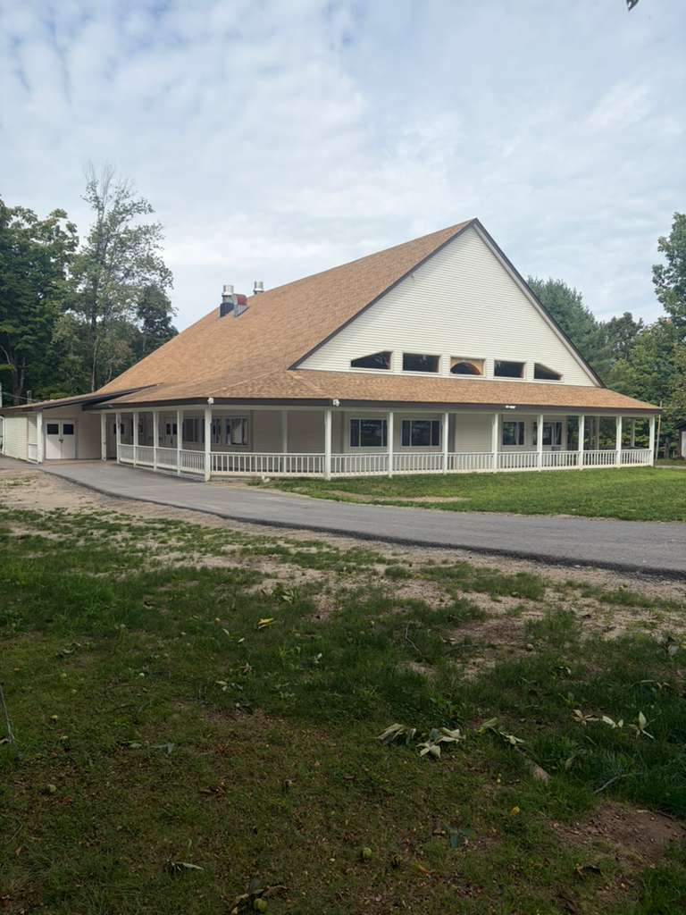 A shul seen from the drive, a steep hipped roof coming down over a covered porch that wraps the building, with clerestory windows set into the gable