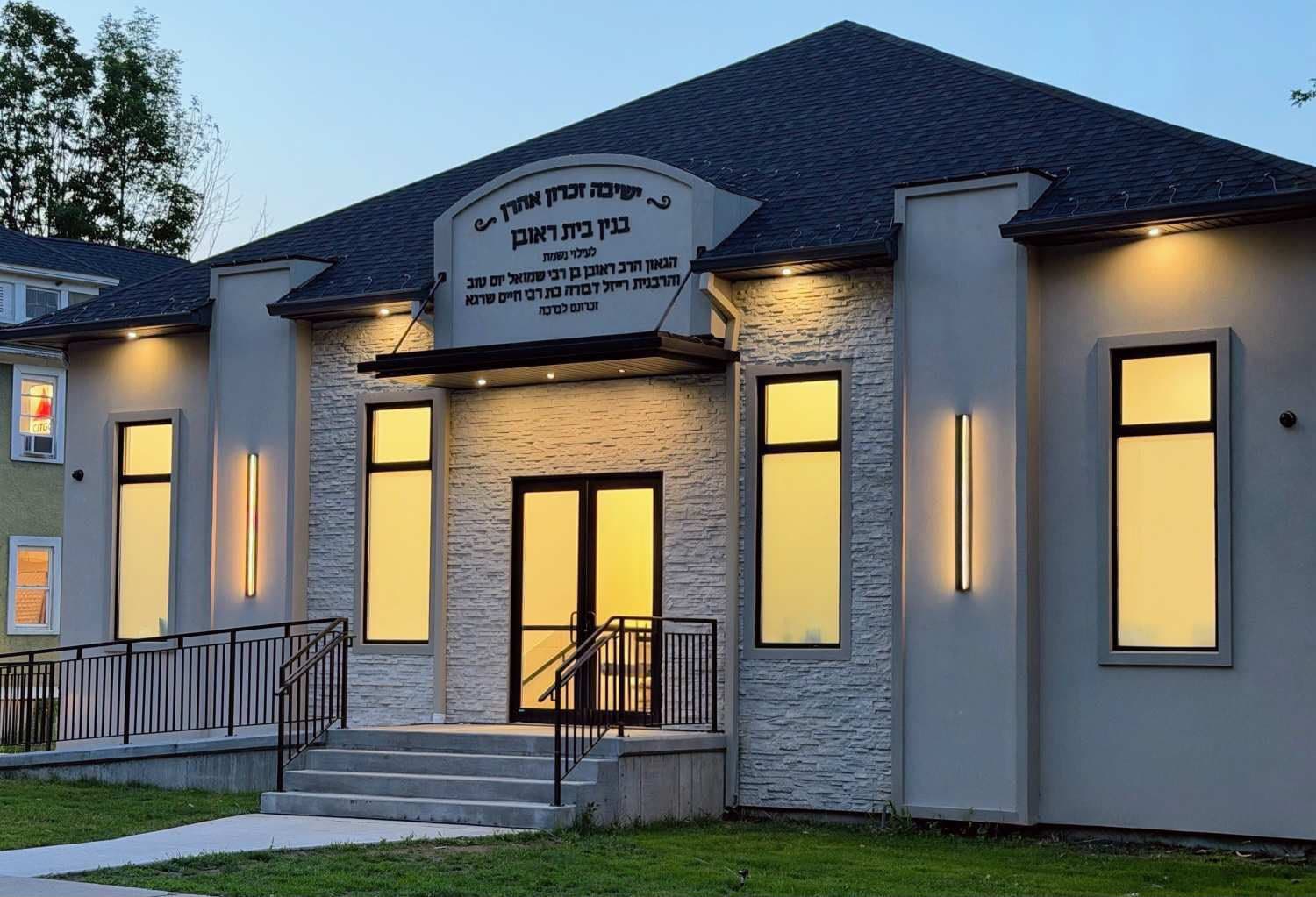 A new beis medrash photographed at dusk — stone and render facade, tall windows lit from within, and a carved dedication panel above the entrance