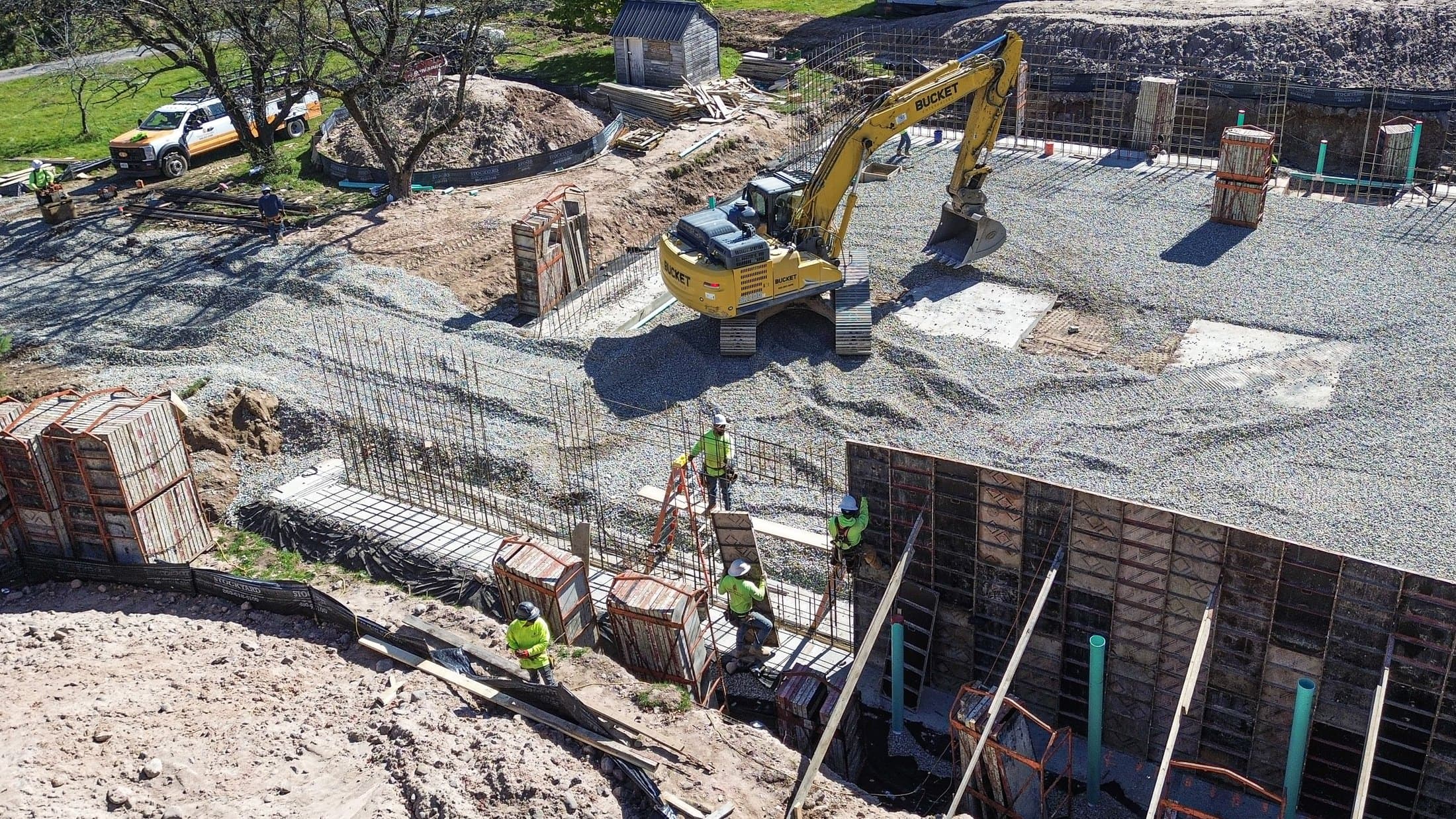 An excavator spreading stone inside a newly poured foundation, formwork and rebar along the near wall
