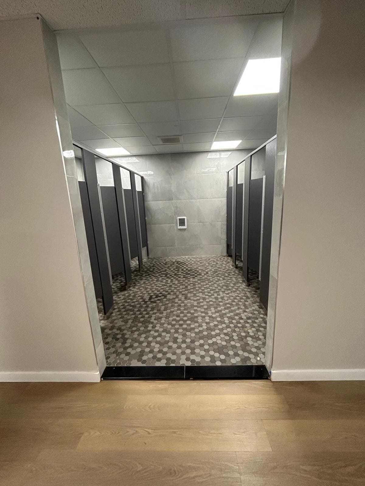 A washroom seen through a mirror-lined opening, cubicles down both sides and a hexagonal mosaic floor