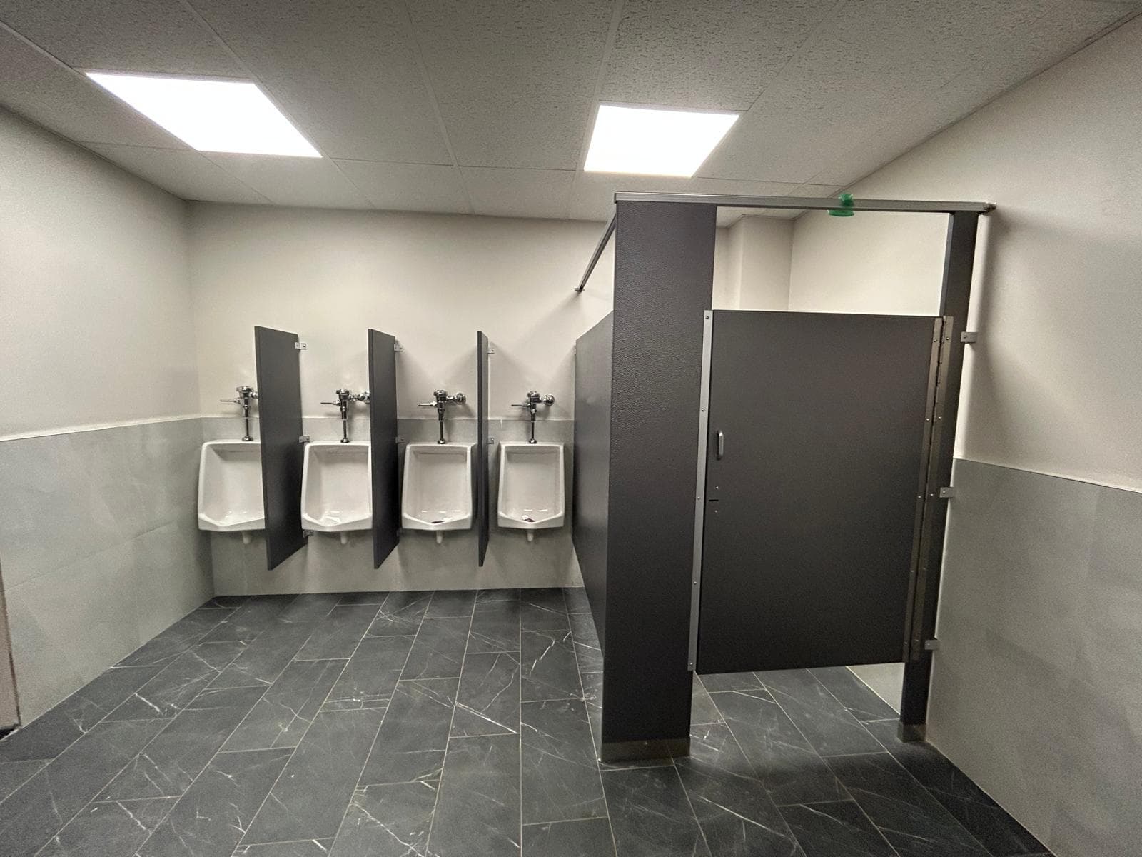 An institutional washroom with four urinals behind privacy screens and an accessible cubicle alongside
