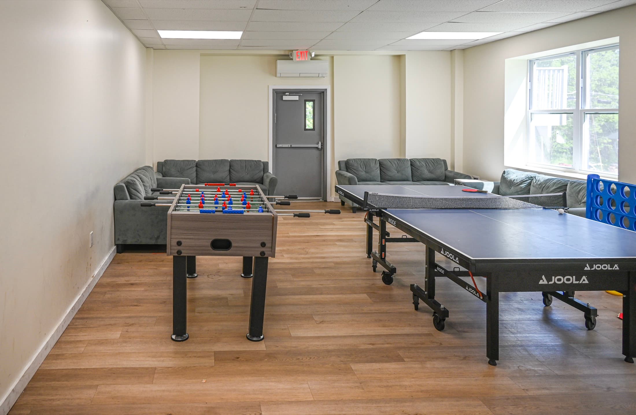 A camp game room with a ping-pong table, foosball table and seating along the window wall